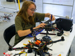 A woman seated at a bench works on two drones